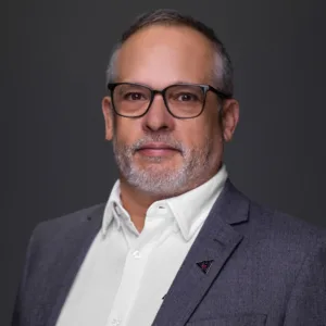 A middle-aged man with glasses and a beard, wearing a gray blazer over a white shirt, poses against a dark background.