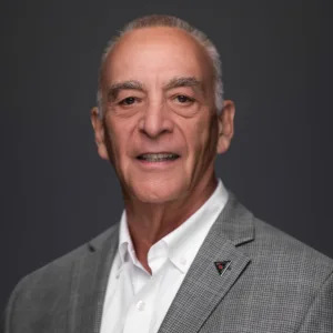 A smiling older man with gray hair, wearing a gray blazer and white shirt, against a dark background.
