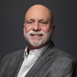 A smiling man with a bald head and white beard, wearing a gray blazer and white shirt, against a dark background.