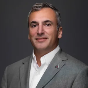 A man with short gray hair, wearing a gray blazer over a white shirt, smiles against a dark background.