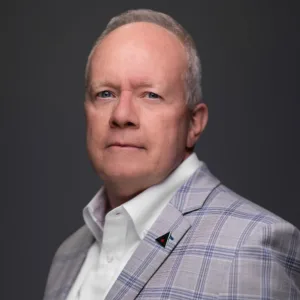 A man with short gray hair, wearing a light plaid blazer and a white shirt, poses against a dark background.